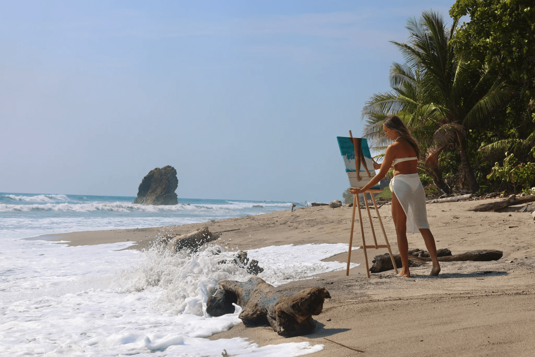 Mariel painting at an easel on the beach in Santa Teresa, with the iconic Playa Santa Teresa rock formation in the background